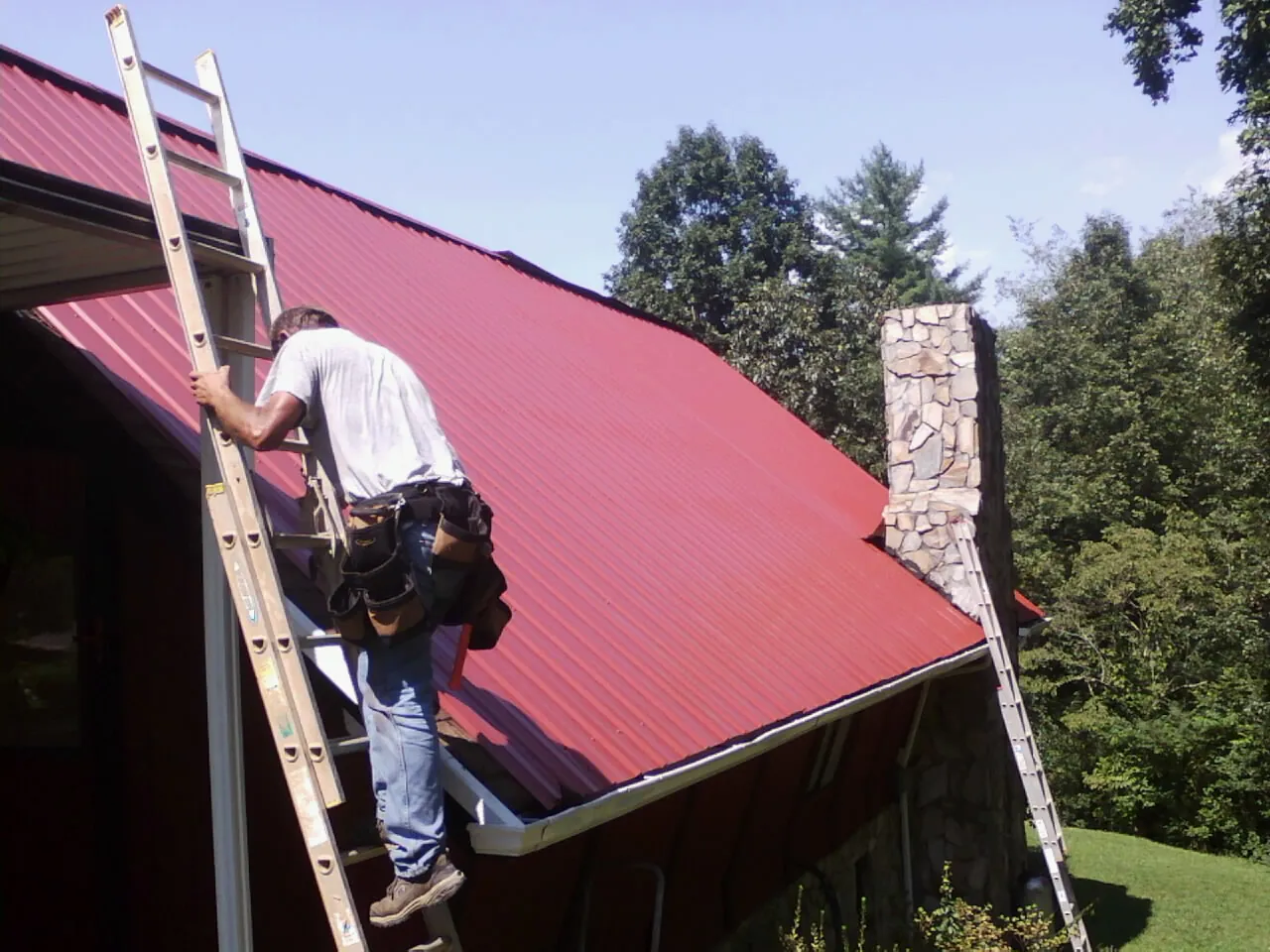 Roofer on a ladder beside a red metal roof and stone chimney — Soffit & Fascia Repair by Roofing Brothers in Silt